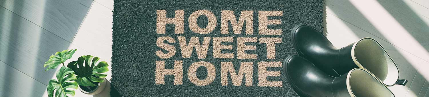Door mat on the floor with the words, 'HOME SWEET HOME' printed on it, a pair of wellingtons and a houseplant.